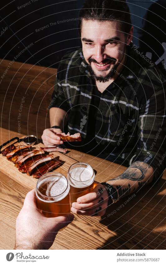 Freundlicher Mann genießt Rippchen und Bier in einem Grillrestaurant Tattoo Vollbart Lächeln Barbecue Restaurant Rippen speisend Teilen Tisch hölzern Glas