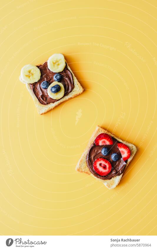 Zwei Toastscheiben mit Schokoladecreme und lustigen Tiergesichter aus Obst. Draufsicht, gelber Hintergrund. Toastbrot schokoladecreme Frühstück Brot lecker