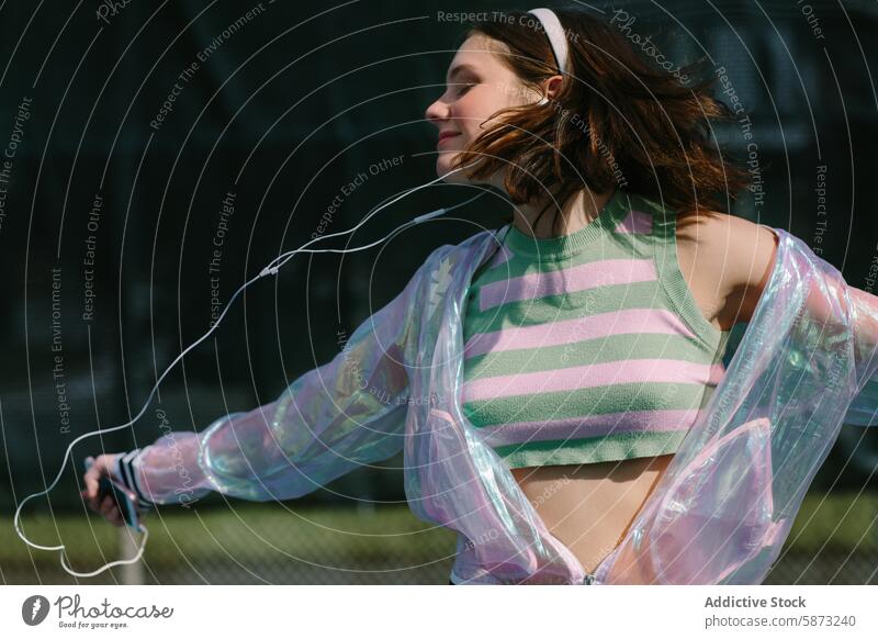 Junge Frau genießt Musik auf einem sonnigen Tennisplatz jung Tag hören Freude Freiheit stylisch transparente Jacke gestreiftes Oberteil pulsierend hell