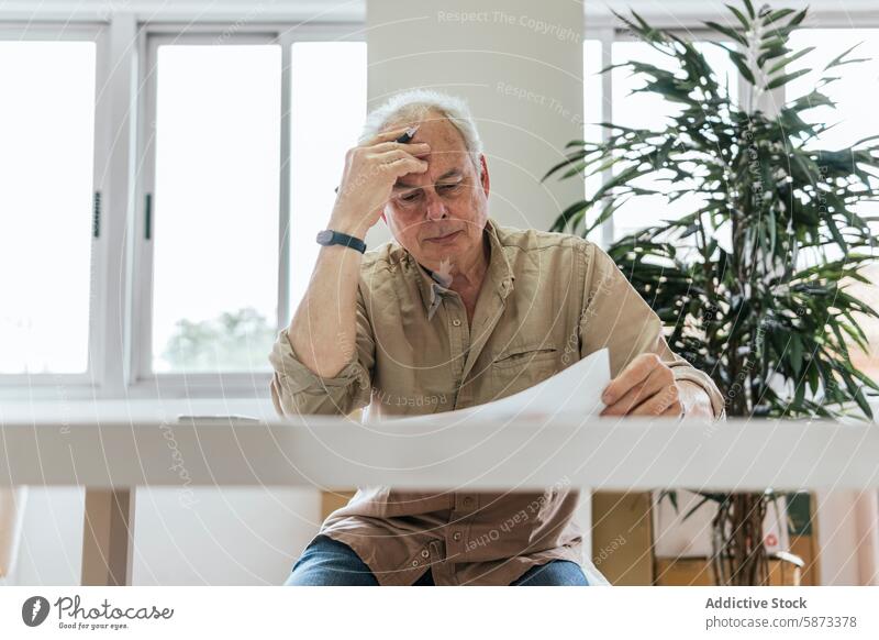 Nachdenklicher älterer Mann, der in einem hellen Raum besorgt auf Papierkram schaut Senior Kaukasier Sorge Stress Tisch weiß gut beleuchtet im Innenbereich