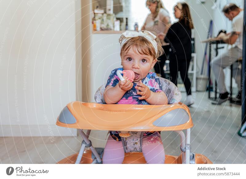 Kleinkind genießt eine Mahlzeit im Hochstuhl, während die Familie im Hintergrund beschäftigt ist Küche heimwärts im Innenbereich Baby Mädchen Spielzeug lässig