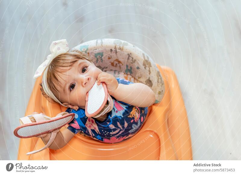 Baby-Mädchen kaut auf ihrem Schuh, während sie in einem Hochstuhl sitzt heimwärts spielerisch niedlich Kind Säugling Kleinkind Familie sitzen Schleife Stirnband