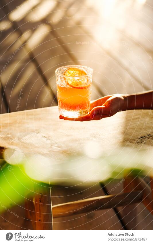 Eine unkenntliche Hand, die einen erfrischenden Cocktail an einem sonnenbeschienenen Tisch im Freien hält Sonnenlicht Sommer Erfrischung Orangenscheibe