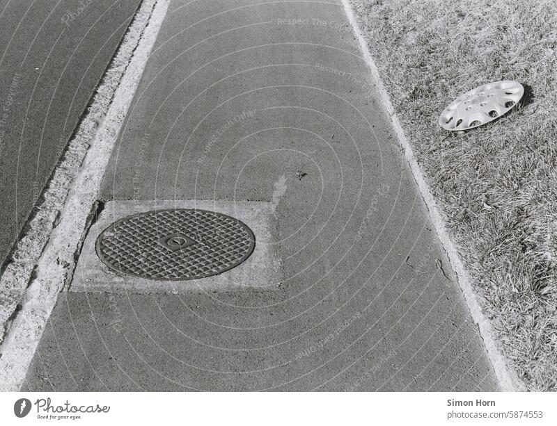 Gehweg mit Gullideckel, daneben liegt eine Radkappe auf einem Rasenstreifen Fußweg Spaziergang Weg rund Asphalt Schwarzweißfoto Schwarz-Weiß-Fotografie