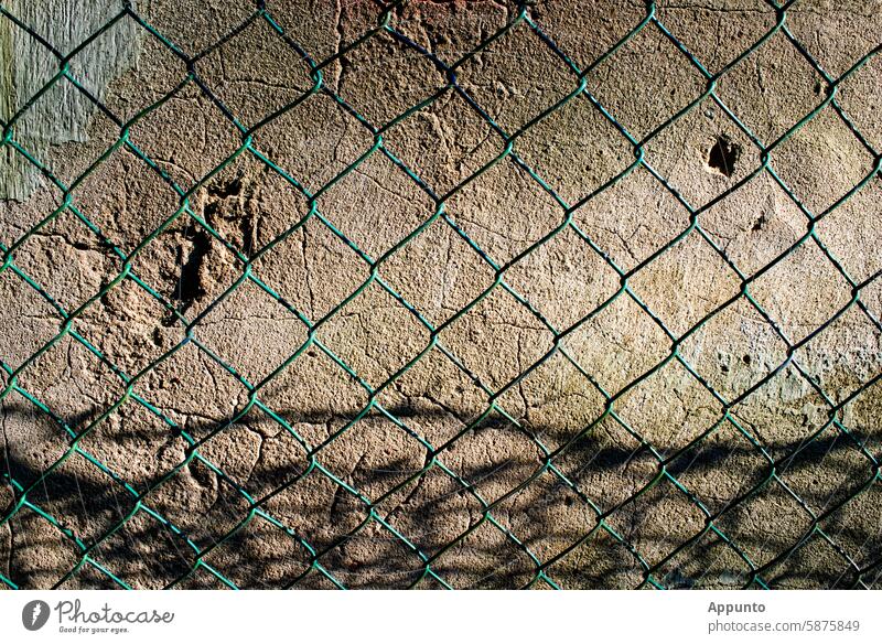 Wand, Zaun und Zeit - Details einer alten Wand mit Rissen und narbenartigen Vertiefungen hinter einem schattenwerfenden grünen Maschendrahtzaun Mauer Zement