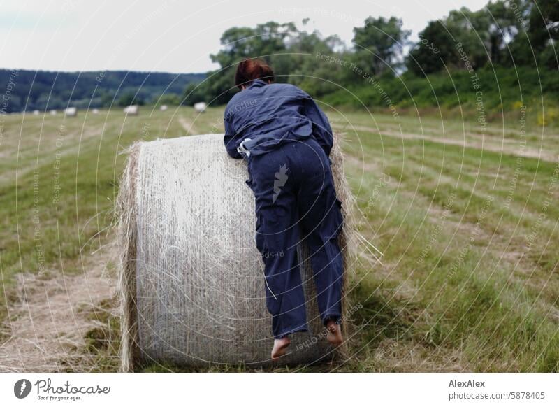 Eine Frau in einer Art Blaumann versucht barfuß auf einen runden Strohballen zu klettern, man sieht sie nur von hinten Landschaft bafuß Feld Sommer Gras Ernte
