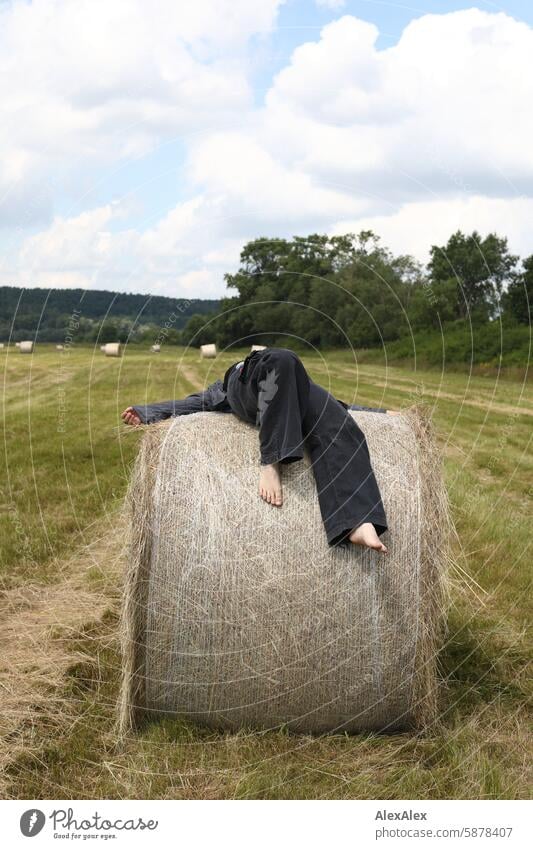 Eine Frau in einer Art Blaumann ist barfuß auf einen runden Strohballen geklettert und liegt nun oben auf Landschaft bafuß Feld Sommer Gras Ernte Landwirtschaft