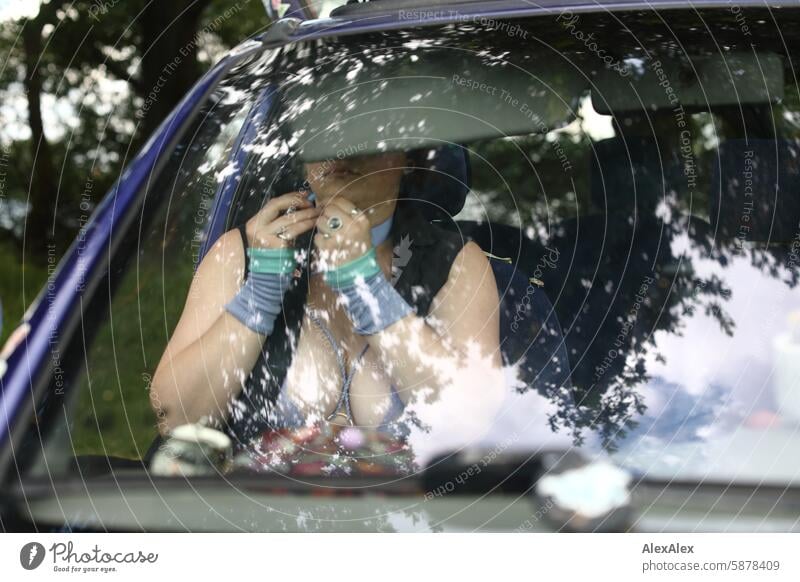 Eine Frau in einer mit großem Ausschnitt sitzt in ihrem Auto auf der rechten Seite vorne und schminkt sich, legt Schmuck an. Man kann sie durch die Reflexion in der Windschutzscheibe nicht erkennen