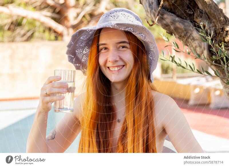 Junge Frau genießt einen sonnigen Sommertag am Pool Tag Hut Wasser Glas Lächeln heiter im Freien Lifestyle erfrischend entspannt Beckenrand jung Rotschopf