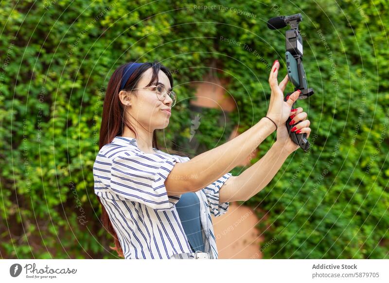 Junge Frau beim Vloggen im Freien mit einem mobilen Gimbal Vlogging Mobile Kardangelenk Smartphone Video Aufnahme Garten üppig (Wuchs) grün