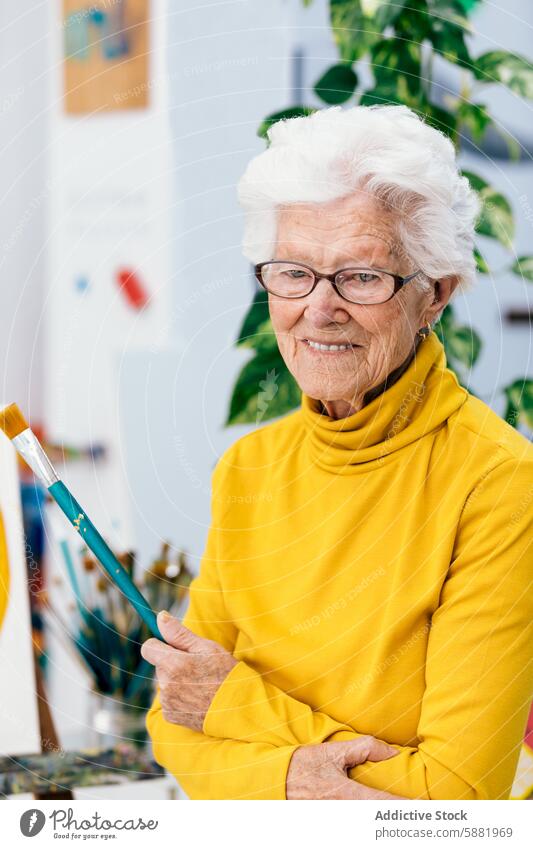 Ältere Künstlerin hält einen Pinsel in einem Atelier Frau älter Senior reif Malerei heiter gelb Rollkragenpulli Pinselblume Kunst Proviant Kreativität Hobby