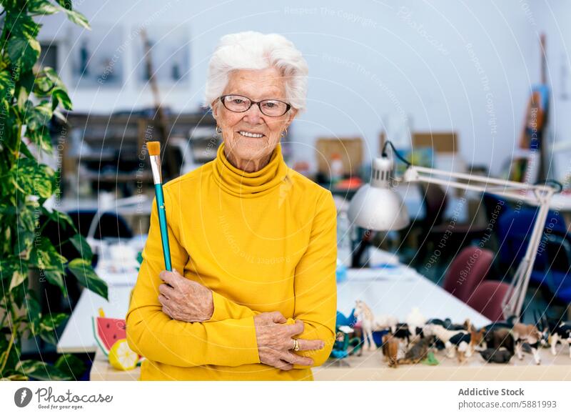 Ältere Künstlerin hält einen Pinsel im Atelier älter Frau Pinselblume heiter Senior reif weiße Haare Lächeln gelb Rollkragenpulli Kunst die Arme verschränkt