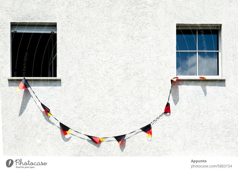 Keep smiling! - Zur Fußball-Europameisterschaft 2024 ist an einer weißen Hausfassade zwischen zwei Fenstern eine Leine mit schwarz-rot-goldenen Deutschland-Wimpeln aufgespannt, so dass der Eindruck eines zwinkernden Smileys entsteht.