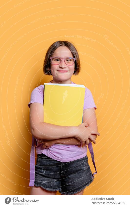 Junges Mädchen mit Brille hält ein Buch in einem Atelier Lächeln gelber Hintergrund lila t-shirt Jeans-Shorts Kind Bildung studierend Glück lesen Schulkind Frau