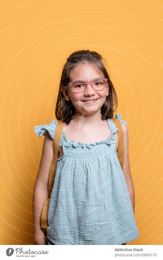 Lächelndes Schulmädchen mit Brille vor einem gelben Hintergrund Mädchen gelber Hintergrund blaues Kleid Porträt Atelier Kind Frau Bildung heiter pulsierend