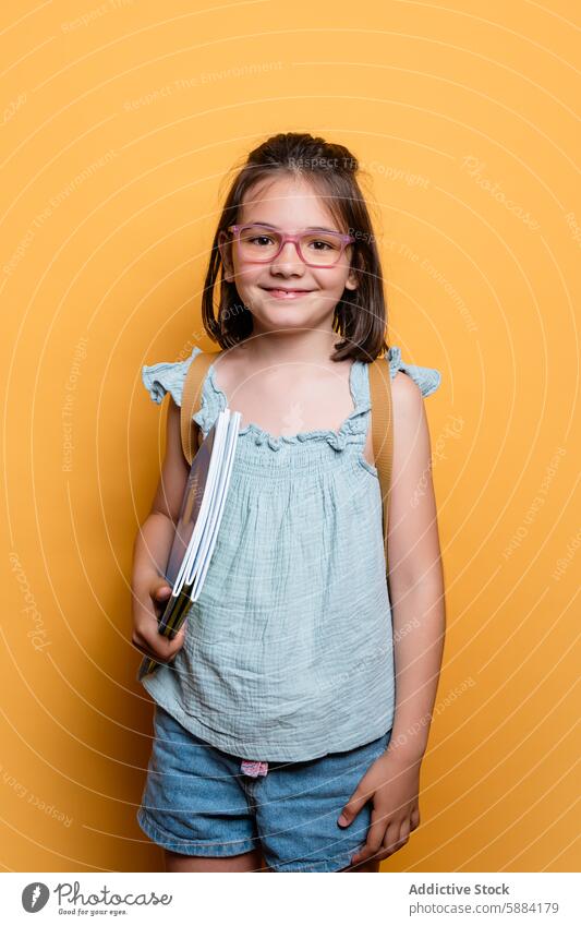 Mädchen im Schulalter mit Büchern lächelnd vor gelbem Hintergrund Schule Lächeln Brille Jeansstoff Atelier Kind Bildung Gelehrter Pupille Fröhlichkeit Mode