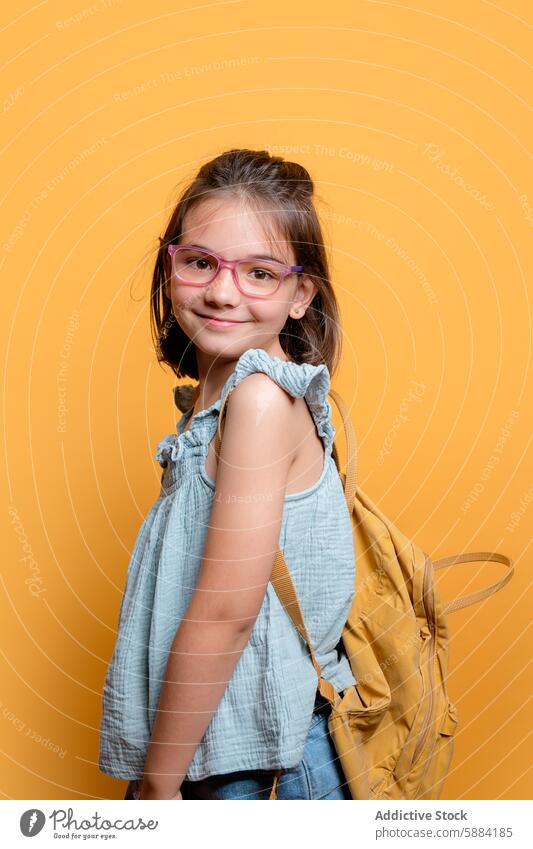 Junge Schülerin mit Rucksack lächelnd auf gelbem Hintergrund Mädchen Schule Lächeln Brille Kind Atelier Porträt Glück heiter Mode Accessoire gerüschtes Oberteil