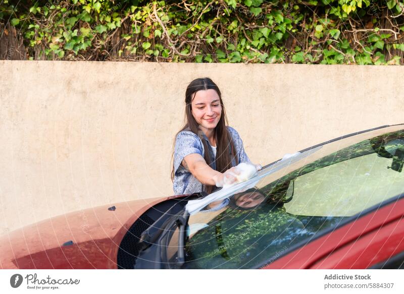 Junge Frau wäscht die Windschutzscheibe eines Autos im Freien Autowäsche Reinigen Lächeln Seife Wasser Rakel Fahrzeug Autopflege manuell Wehen Hobby sonnig Tag