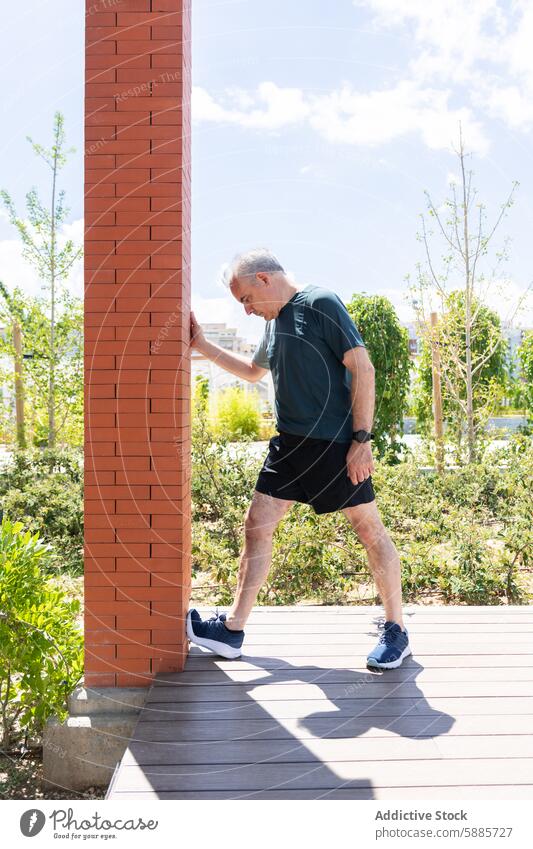 Älterer Mann, der sich in einem sonnigen Park gegen eine Mauer lehnt Senior reif strecken Sportbekleidung im Freien Bein Baustein Spalte Baum blau Himmel