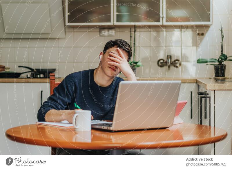 Frustrierter junger Mann arbeitet von zu Hause aus an einem Laptop Stress überwältigt Fernarbeit Küche Tisch Kaffeebecher Papier Heimarbeitsplatz Frustration