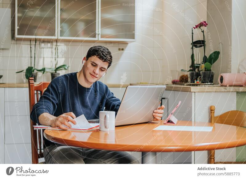 Junger Mann beim Lernen mit digitalen Geräten in der heimischen Küche jung studierend heimwärts Laptop Notebook Smartphone Tisch Stuhl männlich Erwachsener