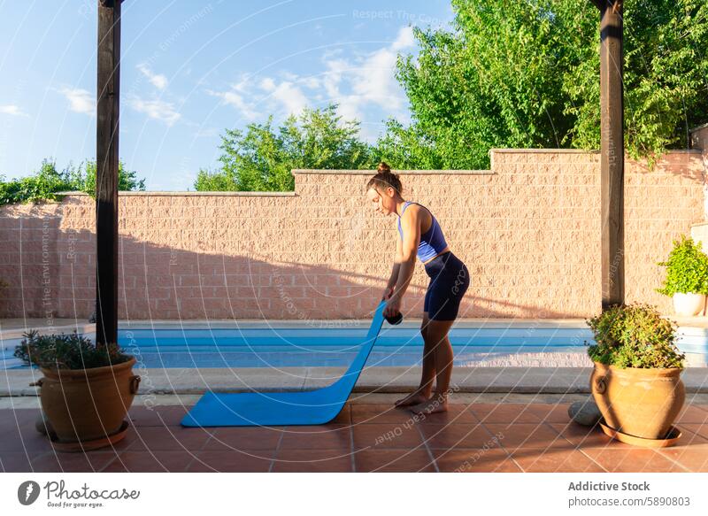 Frau bereitet sich auf eine Yoga-Sitzung am Pool vor im Freien Fitness Übung Gesundheit Wellness ruhig sonnig Hinterhof Unterlage abrollen vorbereitend Kleidung