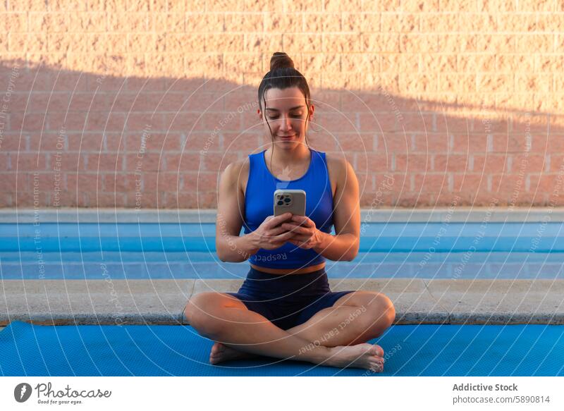 Junge Frau mit einem Mobiltelefon auf einer Yogamatte neben einem ruhigen Schwimmbad bei Sonnenuntergang Pool Smartphone Fitness Gesundheit Gelassenheit