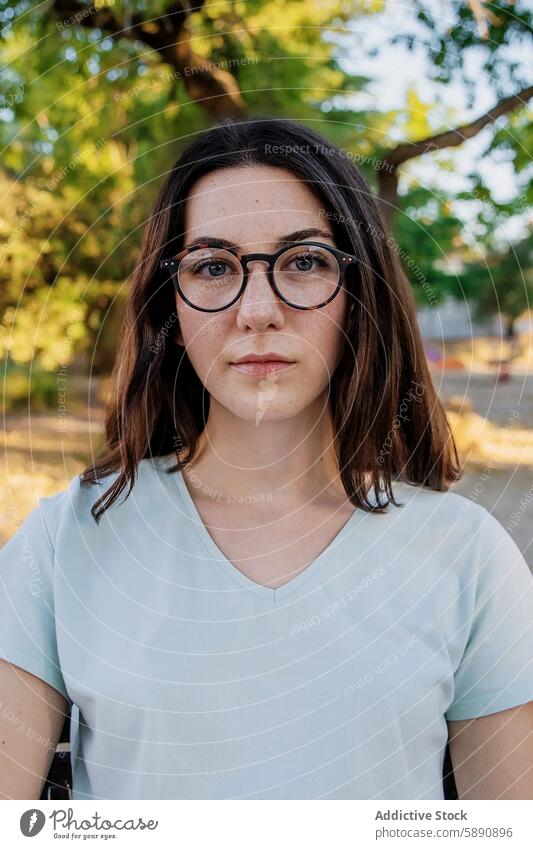 Junge Frau mit Brille schaut in die Kamera in einem Park jung in die Kamera schauen Porträt Kaukasier hell-blau T-Shirt neutrale Meinungsäußerung Baum