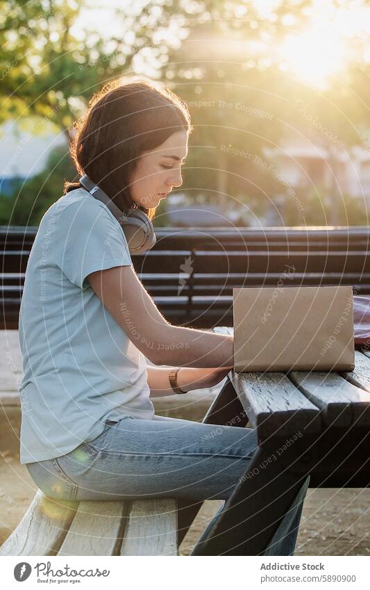 Junge Frau schreibt auf einer Parkbank in ein Notizbuch Bank schreibend Notebook Kopfhörer Konzentration hintergrundbeleuchtet Sitzen herabsehend im Freien