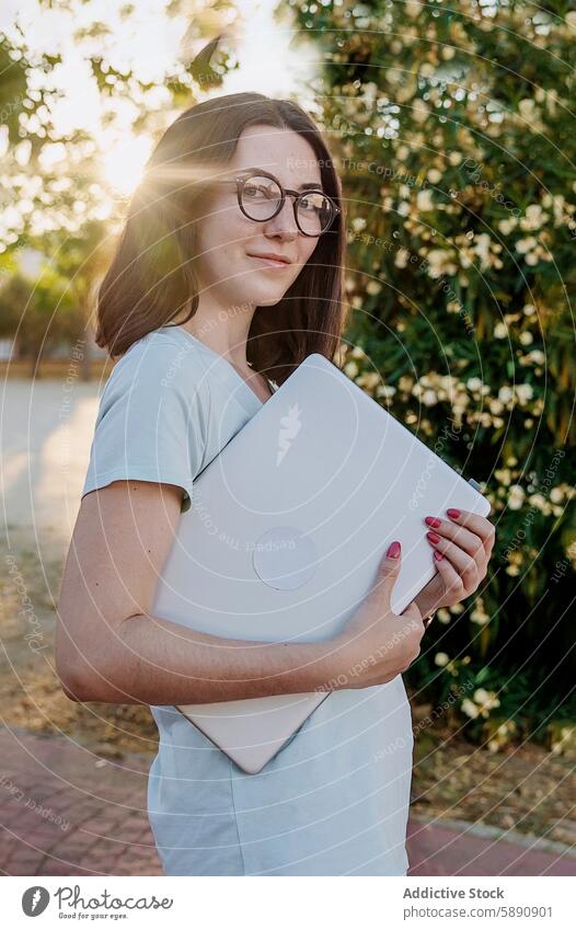 Junge Frau mit Brille hält einen Laptop im Freien Garten Sonnenlicht Technik & Technologie Apparatur Gerät jung Umarmen Halt braune Haare lässig Sommer Tag