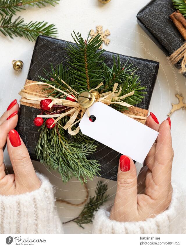 Hände mit Weihnachtsgeschenk mit horizontalem leeren Geschenkanhänger, mit Tannenzweigen und Dekor, Mockup Weihnachten Attrappe präsentieren Postkarte schwarz
