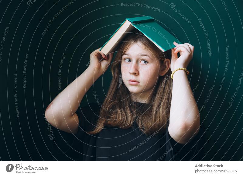 Junges Mädchen mit Buch auf dem Kopf vor dunklem Hintergrund Schüler Bildung zurück zur Schule nachdenklich Porträt Vor dunklem Hintergrund Kontemplation