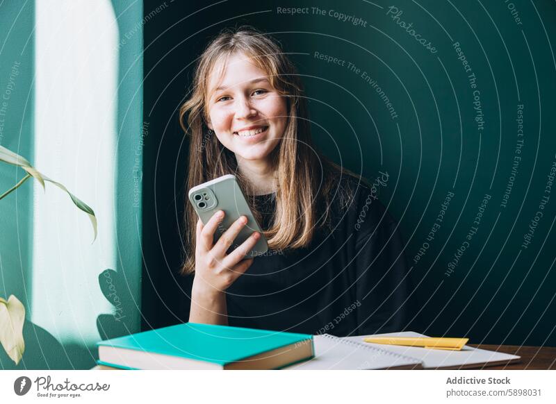 Teenager-Mädchen mit Smartphone am Schreibtisch Handy in die Kamera schauen Lächeln lernen Buch Notebook Schreibstift Bildung Schule Technik & Technologie