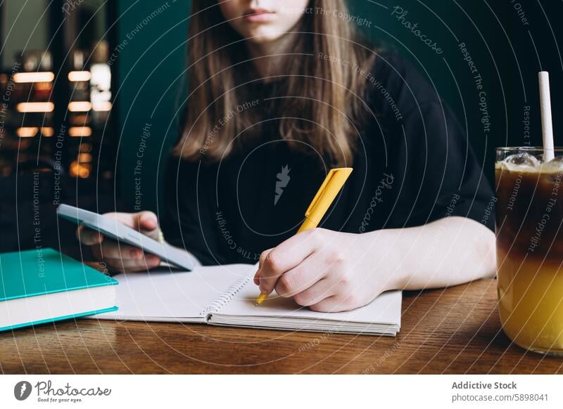 Eine Studentin benutzt ihr Smartphone, während sie in einem Café Notizen macht Schüler Frau Notebook schreibend Schreibstift Tisch trinken kaltes Getränk Glas