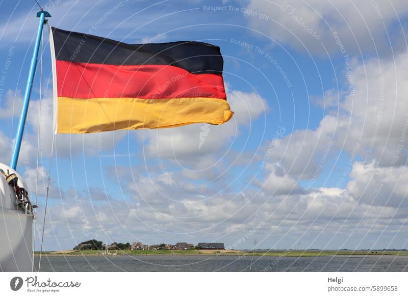 wehende Deutschlandflagge vor blauem Himmel mit Wolken, im Hintergrund die Hallig Oland Flagge Meer Wasser Nordsee Natur Sommer schönes Wetter Außenaufnahme