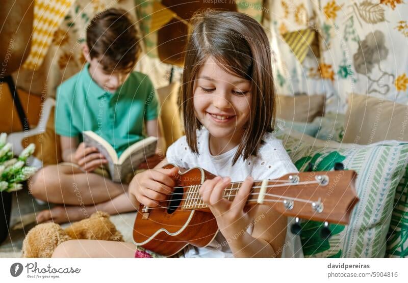 Glücklich lächelndes kleines Mädchen spielt Ukulele, während ein Junge auf einem handgefertigten Schutzzelt ein Buch liest Lächeln Spielen Gitarre lesen