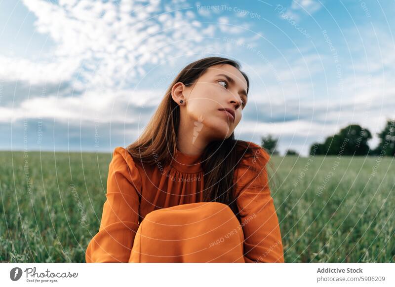 Junge brünette Frau, die in einem grasbewachsenen Feld wegschaut Himmel Wolken Gelassenheit nachdenklich Wegsehen Gras im Freien Natur grün orangefarbene Bluse