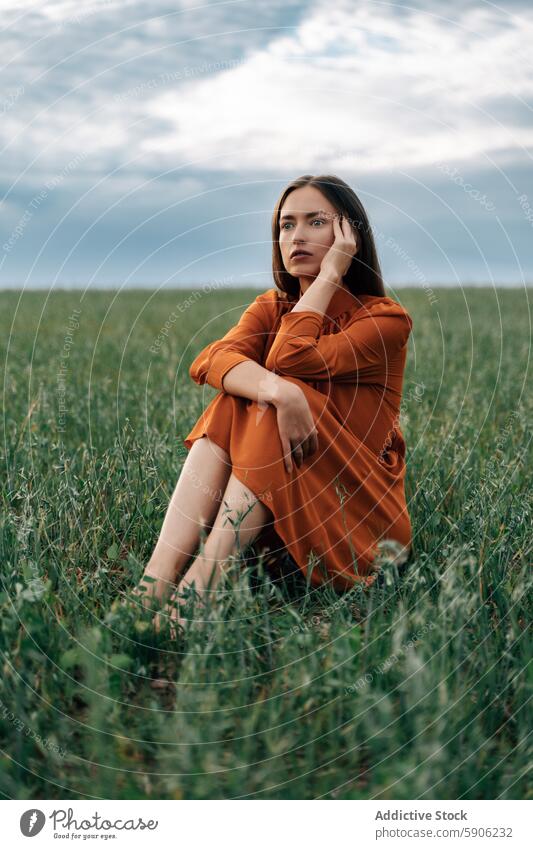 Nachdenkliche brünette Frau sitzt auf einer grünen Wiese Feld orangefarbenes Kleid Sitzen Gras besinnlich nachdenklich beschaulich Natur im Freien Sommer