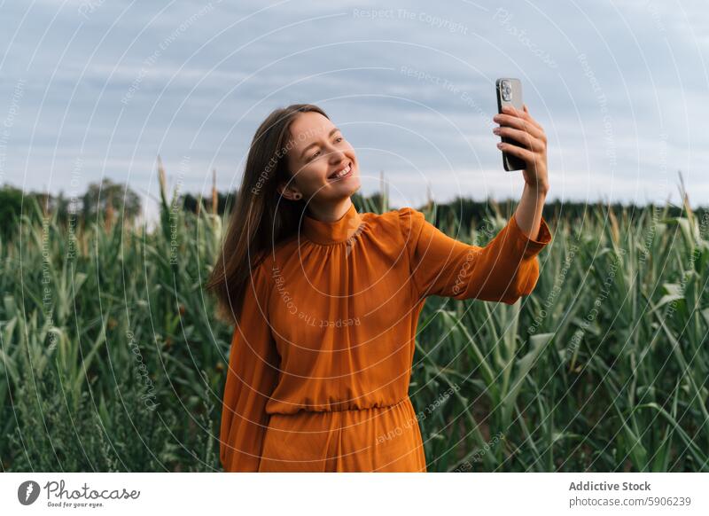 Junge brünette Frau nimmt Selfie in einem grünen Kornfeld Smartphone orangefarbenes Kleid Freude im Freien Handy Natur Ackerbau Sommer lässig Freizeit Gerät