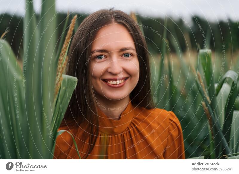 Junge brünette Frau schaut in die Kamera auf einer grünen Wiese Feld orangefarbene Bluse Lächeln in die Kamera schauen Laubwerk Natur im Freien jung heiter