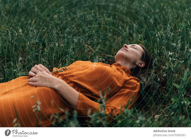 Frau in orangefarbenem Kleid entspannt sich im grünen Feld entspannend brünett Natur Augen geschlossen Ruhe Gras im Freien Sommer Gelassenheit friedlich Wiese