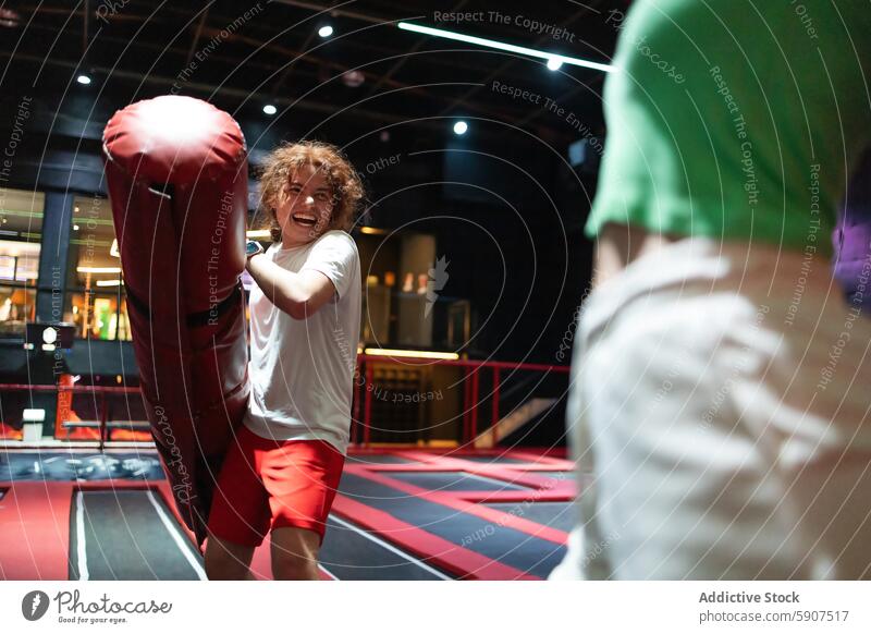 Fröhlicher Mann spielt mit Boxsack im Jumping Club Springclub im Innenbereich lachen Spaß krause Haare energetisch spielerisch rot weißes Hemd rote Shorts