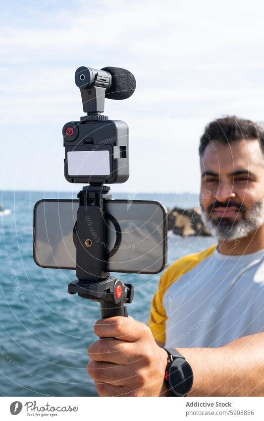 Mann, der mit modernen Geräten ein Video am Meer aufnimmt Inder Sommer Sport Aufnahme Seeküste Smartphone Apparatur Fotokamera Stabilisator