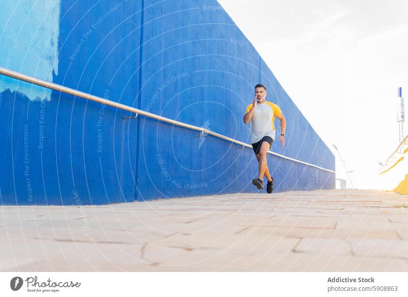 Indischer Mann beim Joggen in einer sommerlichen Umgebung vor einer blauen Wand Inder Jogger rennen Sportbekleidung Sommer im Freien sonnig blaue Wand Aktivität