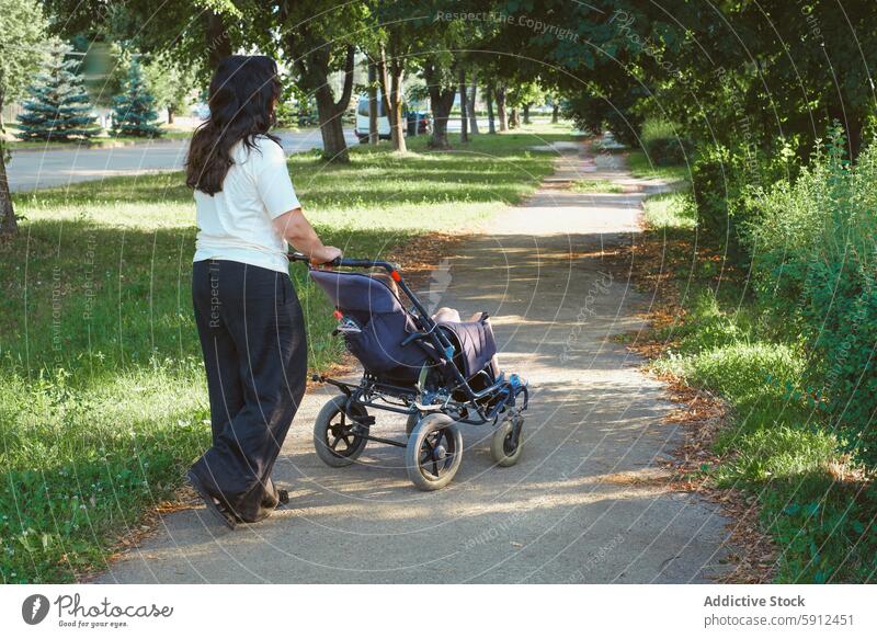 Mutter geht mit Sohn im Rollstuhl im Park spazieren zerebrale Kinderlähmung laufen Natur Weg unkenntlich anonym gesichtslos Rückansicht Baum im Freien Betreuung