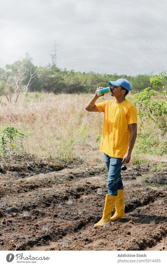 Junger Landwirt tränkt beim Verpflanzen von Hibiskustrieben jung Hydratisierung Verpflanzung Hawaiiblume schießen gelb Hemd blau Verschlussdeckel trinken