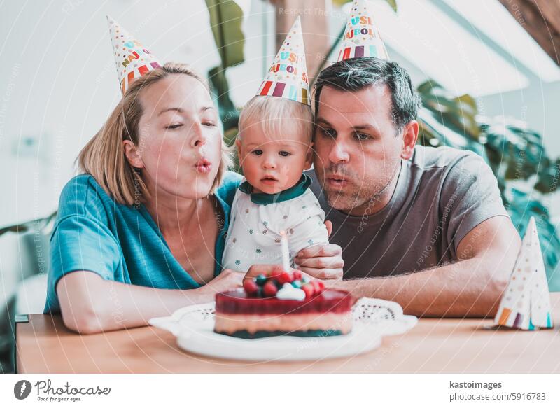 Glücklicher kleiner Junge, der Kerzen auf dem Kuchen ausbläst, während er seinen Geburtstag mit jungen, liebenden Eltern feiert. Das aufgeregte Kind hebt die Arme und lächelt, während es Glückwünsche und Geschenke von der Familie erhält.