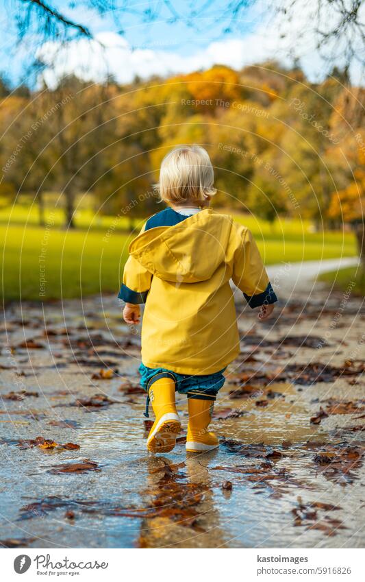 Sonne scheint immer nach dem Regen. Kleine Bindung Kleinkind Junge trägt gelbe Gummistiefel und gelben wasserdichten Regenmantel zu Fuß in Pfützen im Stadtpark auf sonnigen regnerischen Tag.