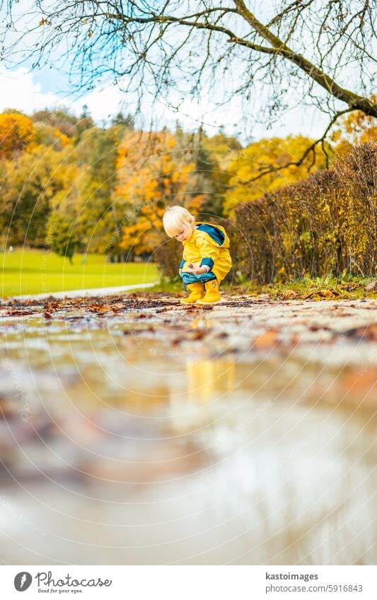 Sonne scheint immer nach dem Regen. Kleine Bindung Kleinkind Junge trägt gelbe Gummistiefel und gelben wasserdichten Regenmantel zu Fuß in Pfützen im Stadtpark auf sonnigen regnerischen Tag.