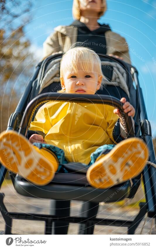 Kleines Kleinkind mit gelbem Regenmantel und Regenstiefeln, das von seiner Mutter im Kinderwagen in einem Stadtpark an einem sonnigen Herbsttag geschoben wird, nachdem der Regen vorbei war.